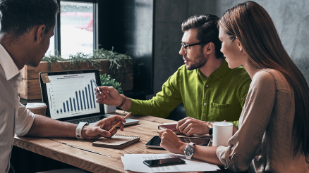 Three BI employees sit together at a table and discuss data analysis on a labtop.